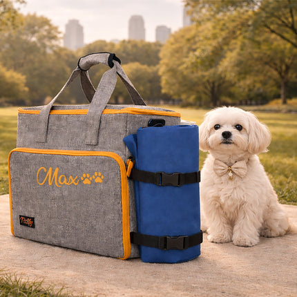 Dog bag with 'Max' and paw print design, blue roll-up mat, and small white dog in a park setting.