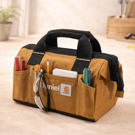Brown tool bag with black handles and Carhartt logo on a light wooden floor.