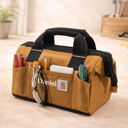 Brown tool bag with black handles and Carhartt logo on a light wooden floor.