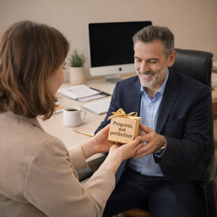 Person handing a gift box with 'Progress, not perfection' message to another person in an office setting.