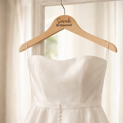 White wedding dress on a wooden hanger with 'Sarah Bridesmaid' engraving, against a neutral background.