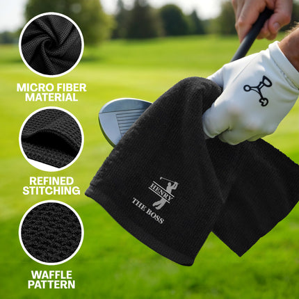 Black golf towel with brand logo held by a gloved hand on a golf course, with text highlighting material and pattern.