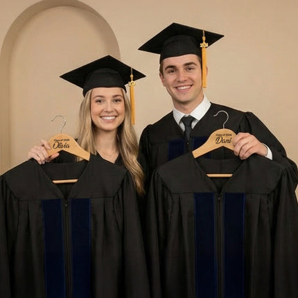 Two graduates in caps and gowns holding hangers with names on them against a beige wall.