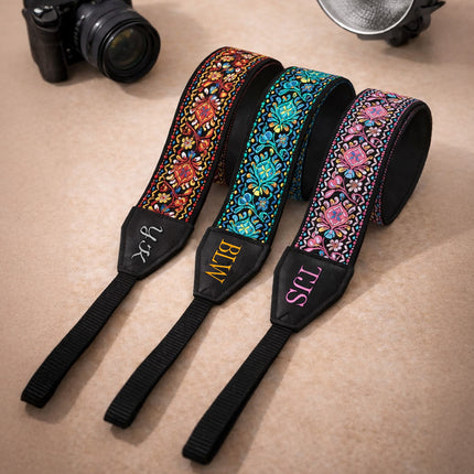 Three colorful camera straps with monograms on a beige surface, with a camera and lens in the background.