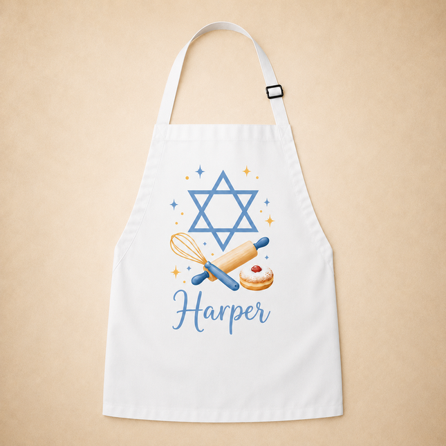 A white apron with a blue Star of David, whisk, rolling pin, donut, and the name 'Harper' printed in blue, displayed against a warm beige background.