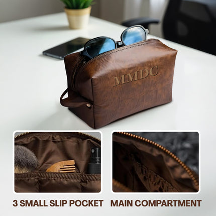 Brown leather toiletry bag with sunglasses on a table, showing interior compartments.