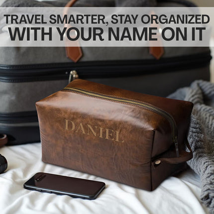 Brown leather toiletry bag with 'DANIEL' engraved, next to a suitcase and phone on a bed.