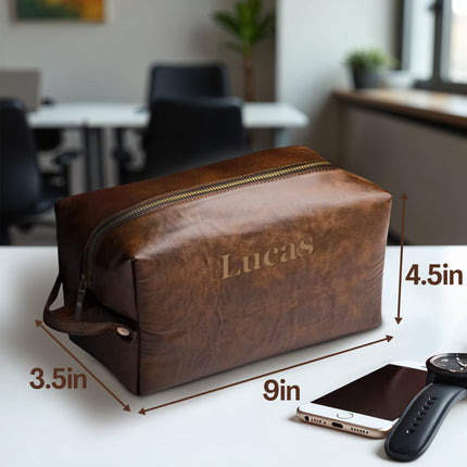 Brown leather toiletry bag with 'Lucas' engraving, shown with dimensions and a smartphone and watch for scale.